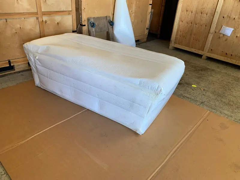 A piece of furniture wrapped in white protective packing on cardboard sheets, beside wooden storage crates.